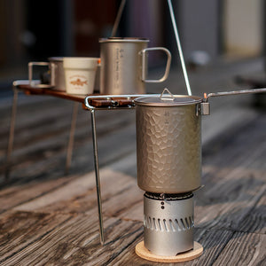 A tall hammered titanium pot on a compact stove, with a mug on a rack behind it.