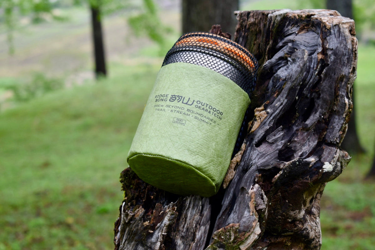 A packed cookware stack in a green sleeve resting against a tree trunk.