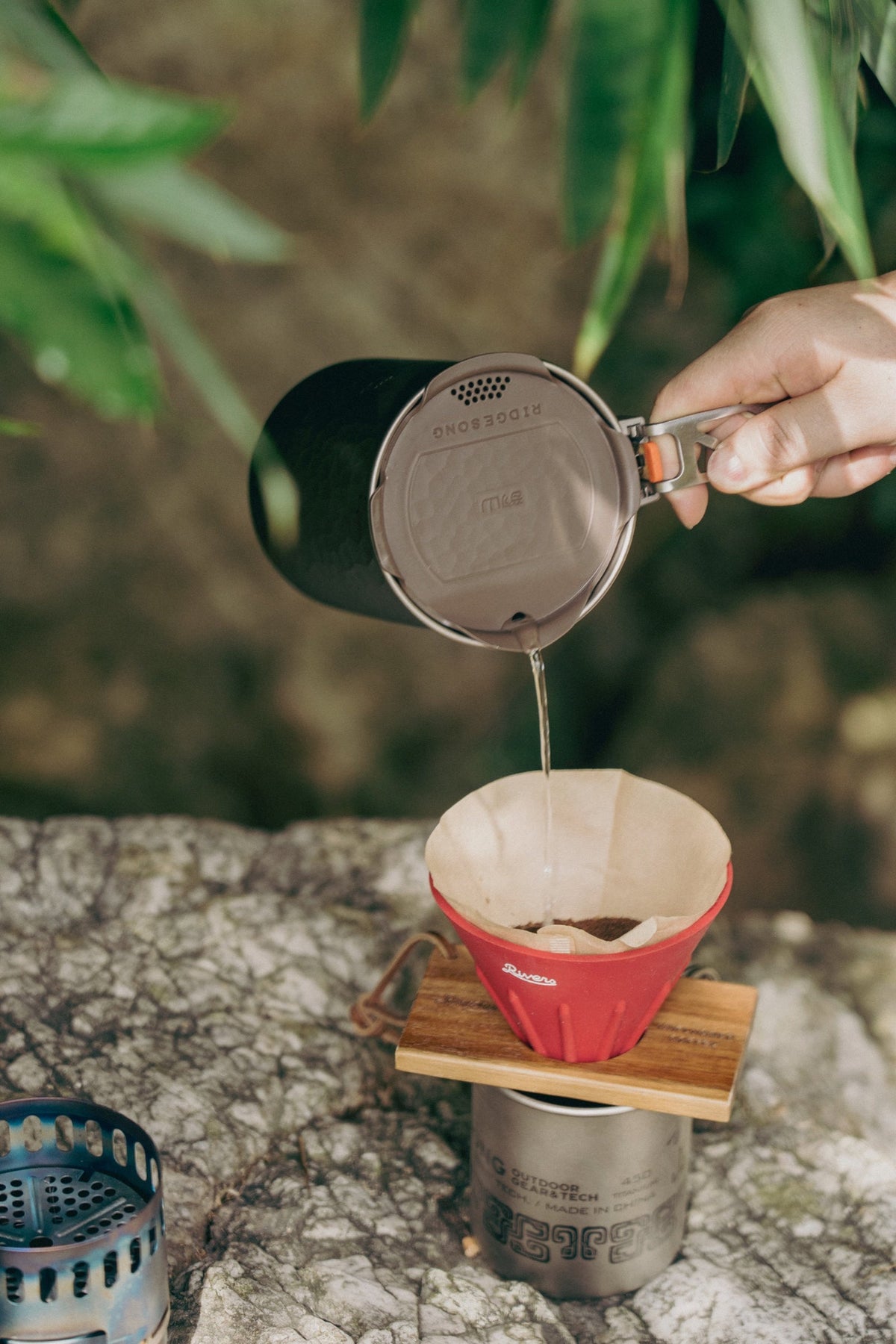 A person pouring water from a titanium pot into a paper-lined red coffee dripper outdoors.